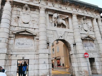 A low-angle photograph of a large, ornate Renaissance stone gate featuring a prominent winged lion statue above the archway, flanked by columns and inscribed plaques, with people walking through the passage.