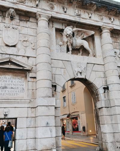 A low-angle photograph of a large, ornate Renaissance stone gate featuring a prominent winged lion statue above the archway, flanked by columns and inscribed plaques, with people walking through the passage.