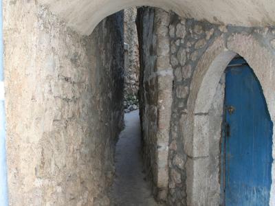 A narrow stone alleyway with a vaulted ceiling, featuring a blue wooden door set into an arched opening on the right side.