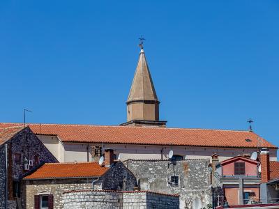 A view of terracotta-roofed stone buildings and a prominent church tower with a pointed spire against a clear blue sky.