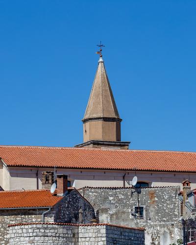 A view of terracotta-roofed stone buildings and a prominent church tower with a pointed spire against a clear blue sky.