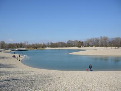 A wide-angle photograph of a large, man-made lake surrounded by extensive pebble beaches under a clear blue sky, with people walking along the shore and a bridge visible in the distance.