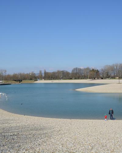 A wide-angle photograph of a large, man-made lake surrounded by extensive pebble beaches under a clear blue sky, with people walking along the shore and a bridge visible in the distance.