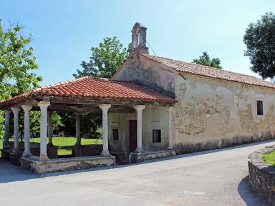 A single-nave stone church with a tiled roof, a bell gable, and an attached open-sided loggia (portico) supported by white columns, situated next to a paved road and green lawn.