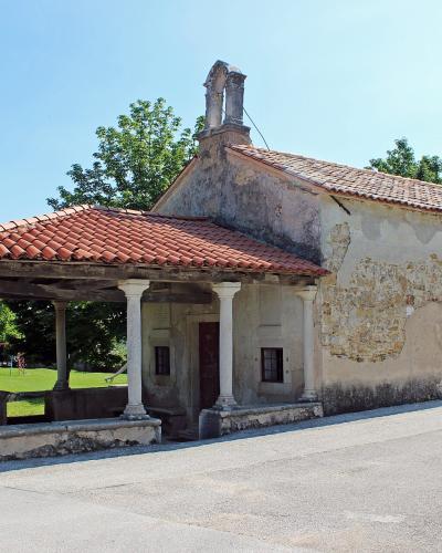 A single-nave stone church with a tiled roof, a bell gable, and an attached open-sided loggia (portico) supported by white columns, situated next to a paved road and green lawn.