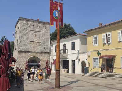 A stone clock tower and a paved town square with a tall wooden mast flying a red banner, surrounded by yellow and white buildings under a clear blue sky.