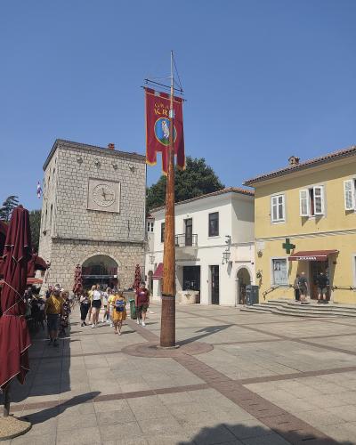 A stone clock tower and a paved town square with a tall wooden mast flying a red banner, surrounded by yellow and white buildings under a clear blue sky.