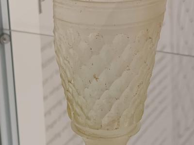 An ancient, faceted glass drinking cup displayed in a museum case with informational text visible on the wall behind it.