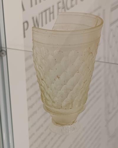 An ancient, faceted glass drinking cup displayed in a museum case with informational text visible on the wall behind it.