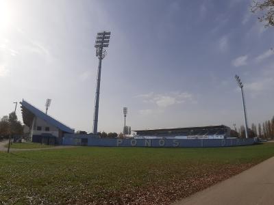 The image shows an outdoor football stadium with blue grandstands, tall floodlight towers, and a grassy field, featuring large white letters spelling "PONOS I DIKA" on a low wall.