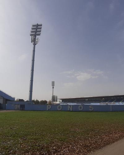The image shows an outdoor football stadium with blue grandstands, tall floodlight towers, and a grassy field, featuring large white letters spelling "PONOS I DIKA" on a low wall.