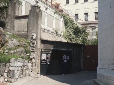 A dark, gated entrance to a tunnel structure set into a rocky hillside, with informational signage on the gate reading "TUNEL" and "ULAZ / ENTRANCE", adjacent to a large white building with barred windows.