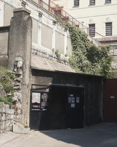 A dark, gated entrance to a tunnel structure set into a rocky hillside, with informational signage on the gate reading "TUNEL" and "ULAZ / ENTRANCE", adjacent to a large white building with barred windows.
