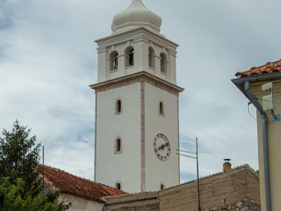 A tall, white bell tower with a clock face, arched belfry openings, and a bulbous dome topped with a cross, rising above surrounding buildings with terracotta roofs.