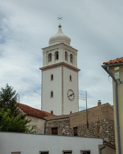 A tall, white bell tower with a clock face, arched belfry openings, and a bulbous dome topped with a cross, rising above surrounding buildings with terracotta roofs.