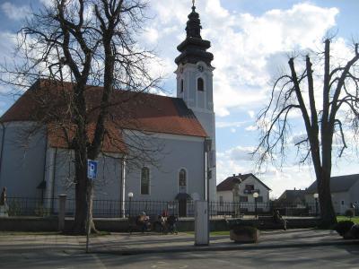 A white baroque-style church with a tall, dark, onion-domed steeple, a terracotta-tiled roof, and a black metal fence in the foreground, with bare trees and people sitting on benches in front.