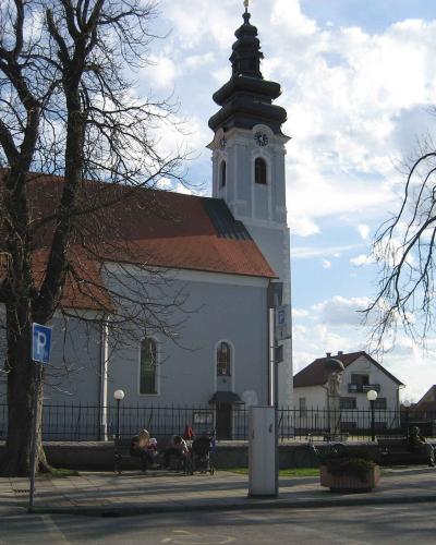 A white baroque-style church with a tall, dark, onion-domed steeple, a terracotta-tiled roof, and a black metal fence in the foreground, with bare trees and people sitting on benches in front.
