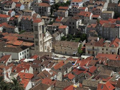An aerial view of a historic Mediterranean town characterized by dense stone buildings with red-tiled roofs, centered around a prominent church with a tall, multi-tiered bell tower and a white facade.