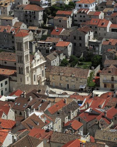 An aerial view of a historic Mediterranean town characterized by dense stone buildings with red-tiled roofs, centered around a prominent church with a tall, multi-tiered bell tower and a white facade.