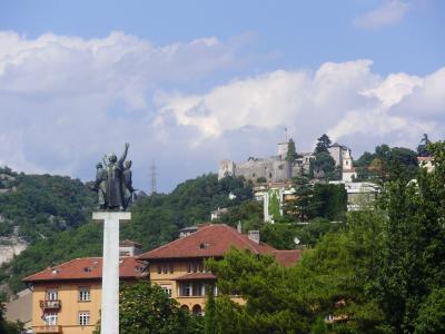 A view of a hilltop castle complex with stone walls and towers, seen from below with a large bronze monument on a white pedestal in the foreground and residential buildings in the mid-ground.