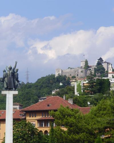 A view of a hilltop castle complex with stone walls and towers, seen from below with a large bronze monument on a white pedestal in the foreground and residential buildings in the mid-ground.