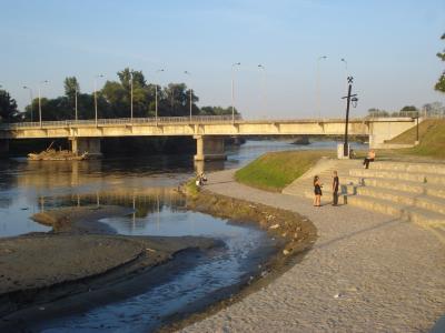 A concrete bridge spanning a river with a cobblestone promenade and stepped seating area along the riverbank, featuring people walking and sitting.