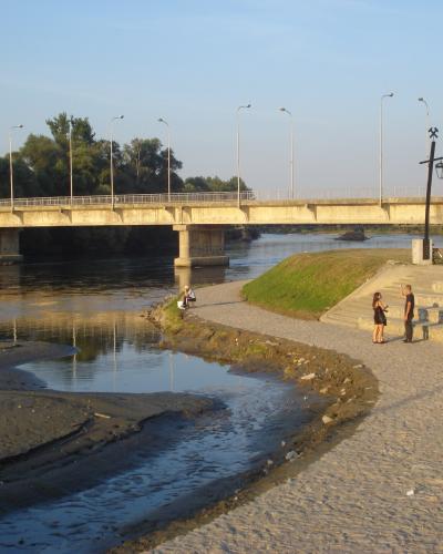 A concrete bridge spanning a river with a cobblestone promenade and stepped seating area along the riverbank, featuring people walking and sitting.