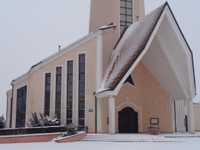 A modern church with a pinkish-beige facade, tall narrow windows, and a large overhanging roof entrance, photographed in snowy winter conditions.