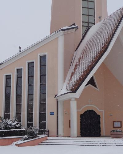 A modern church with a pinkish-beige facade, tall narrow windows, and a large overhanging roof entrance, photographed in snowy winter conditions.