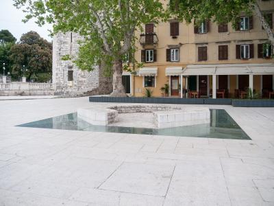 A paved public square featuring a central stone fountain basin surrounded by a shallow glass-covered water feature, with a large stone tower and a yellow building with shops and restaurants in the background.