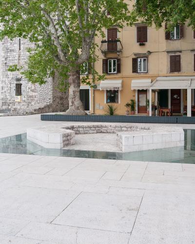 A paved public square featuring a central stone fountain basin surrounded by a shallow glass-covered water feature, with a large stone tower and a yellow building with shops and restaurants in the background.