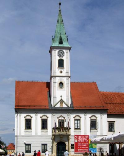 A white Baroque-style town hall building with a tall central clock tower topped by a green copper spire, situated in a paved public square with pedestrians and outdoor cafe seating.