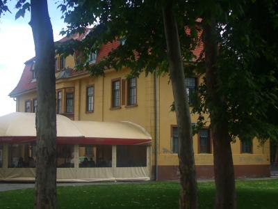 A yellow two-story building with a red-tiled roof, partially obscured by trees, with a large beige outdoor tent structure in front bearing the word "DVORAC" on its awning.