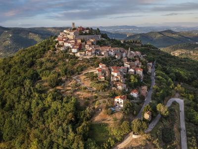 An aerial view of a historic hilltop village with stone buildings, terracotta roofs, and a prominent tower, surrounded by dense green forests and rolling hills.