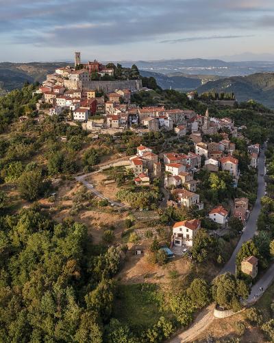 An aerial view of a historic hilltop village with stone buildings, terracotta roofs, and a prominent tower, surrounded by dense green forests and rolling hills.