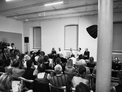 A black and white photograph of an indoor panel discussion or lecture, showing an audience seated in rows facing a stage with five speakers, professional video cameras, and a large lighting umbrella.