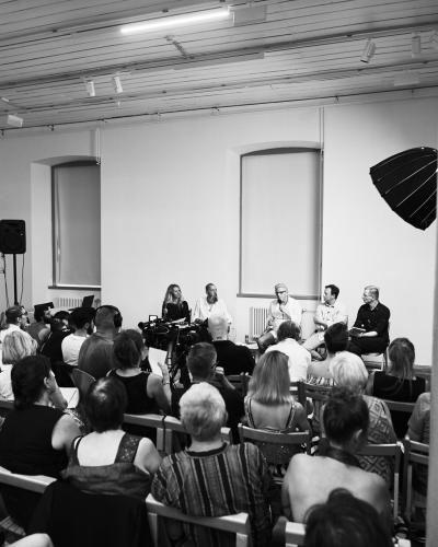 A black and white photograph of an indoor panel discussion or lecture, showing an audience seated in rows facing a stage with five speakers, professional video cameras, and a large lighting umbrella.
