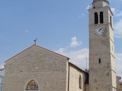 A stone church with a tall, square bell tower featuring a clock and a pointed spire, set against a blue sky.