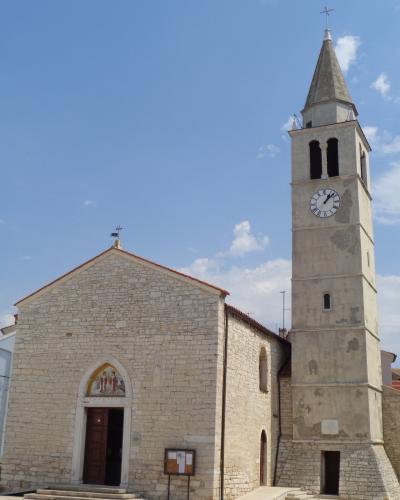 A stone church with a tall, square bell tower featuring a clock and a pointed spire, set against a blue sky.