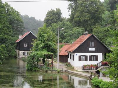 A view of traditional wooden and whitewashed houses along a river in a forested area, with a sign on the white house reading "PENSION VUČETA".