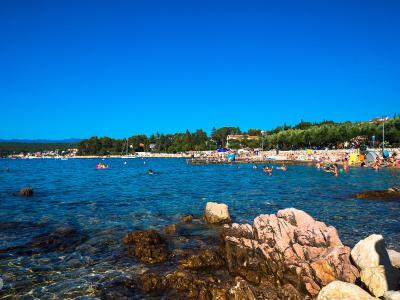 A sunny coastal scene showing a pebble beach crowded with sunbathers and swimmers, with a harbor area and town buildings visible in the background under a clear blue sky.