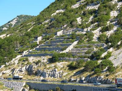A steep, rocky hillside featuring extensive dry-stone terracing, with a highway running along the base where vehicles and a truck with "KONZUM" branding are visible.