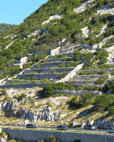 A steep, rocky hillside featuring extensive dry-stone terracing, with a highway running along the base where vehicles and a truck with "KONZUM" branding are visible.
