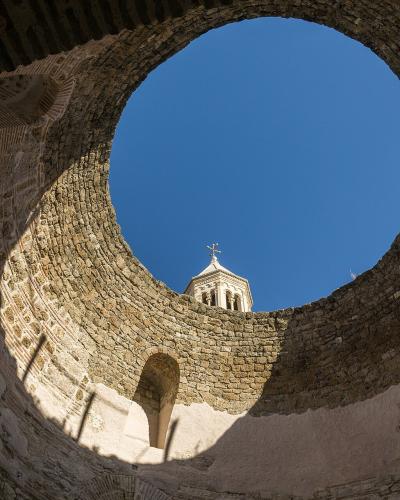 A low-angle view looking up through a circular stone opening (oculus) in an ancient structure, revealing a bell tower with a cross on top against a clear blue sky.