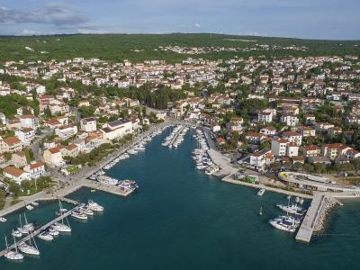 An aerial view of a coastal town with a marina filled with boats, surrounded by white buildings with red-tiled roofs and green hills in the background.