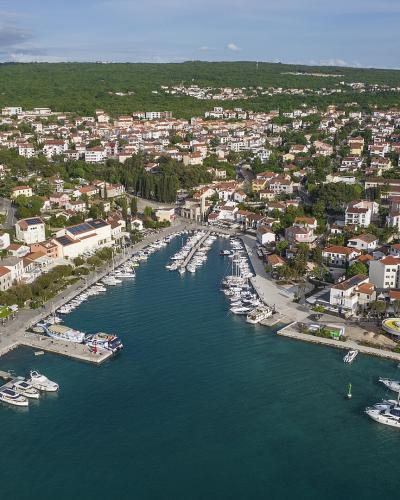 An aerial view of a coastal town with a marina filled with boats, surrounded by white buildings with red-tiled roofs and green hills in the background.