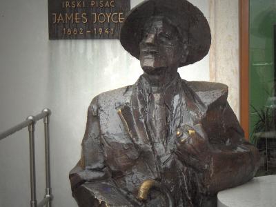 A bronze statue of a seated man wearing a hat and holding a cane, with a plaque behind it reading "IRSKI PISAC JAMES JOYCE 1882-1941", situated next to a white table and metal railing.