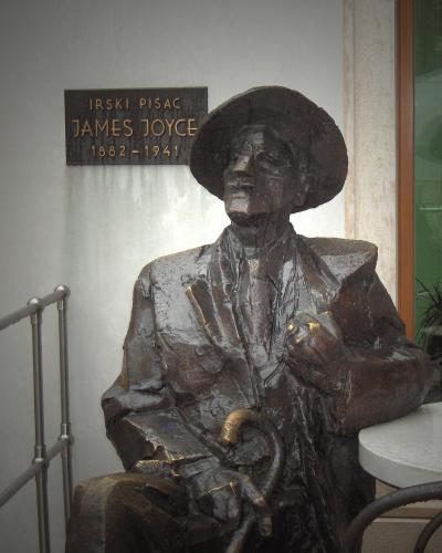A bronze statue of a seated man wearing a hat and holding a cane, with a plaque behind it reading "IRSKI PISAC JAMES JOYCE 1882-1941", situated next to a white table and metal railing.