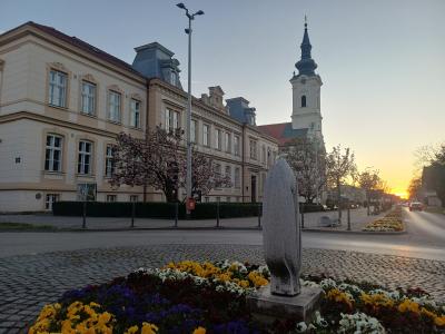 A street scene in a European town square at sunset, featuring a row of multi-story buildings with classical facades, a tall church tower with a dark spire in the background, and a modern abstract stone sculpture in the foreground surrounded by flower