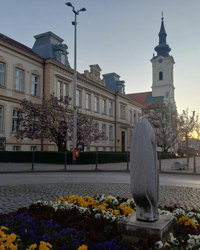 A street scene in a European town square at sunset, featuring a row of multi-story buildings with classical facades, a tall church tower with a dark spire in the background, and a modern abstract stone sculpture in the foreground surrounded by flower